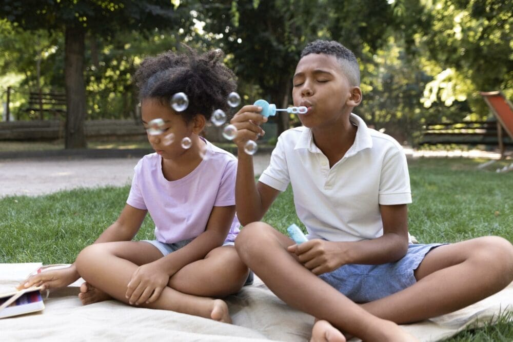 A boy is blowing soap bubbles while sitting on a blanket in a sunny park