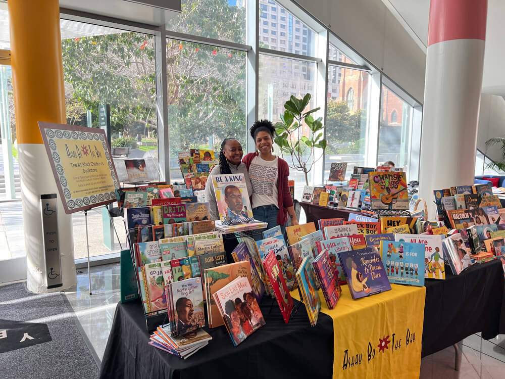 Two people smile at a colorful book display in a bright room with large windows.